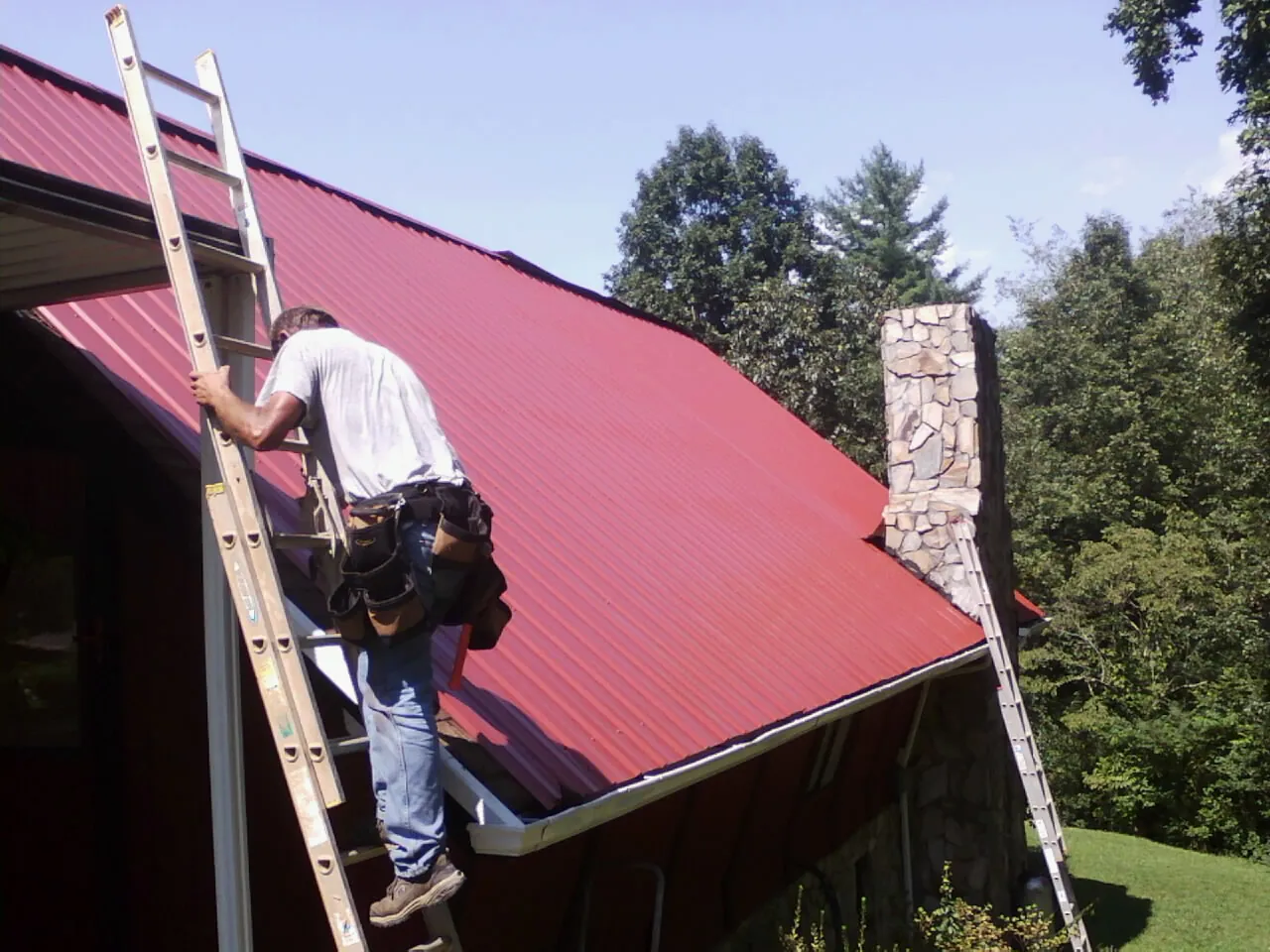 Roofer on a ladder beside a red metal roof and stone chimney — Soffit & Fascia Repair by Roofing Brothers in Medon